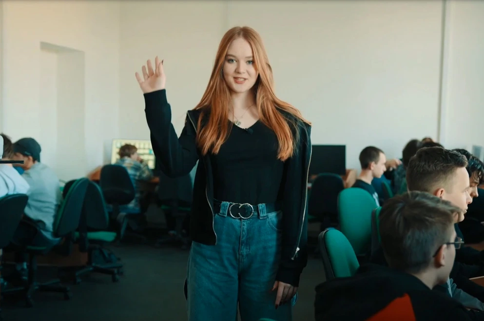 Uczennica uśmiecha się i macha ręką do kamery, witając widzów.